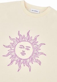 T-shirt en coton de couleur crème avec un design de soleil brodé en violet, positionné au centre, avec des rayons stylisés s'étendant vers l'extérieur.