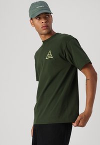 T-shirt vert olive à manches courtes avec un logo triangulaire jaune sur la poitrine, assorti à une casquette vert clair. Jean noir et tatouage sur le bras visible.