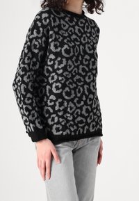 Schwarzer Pullover mit grauem Leopardenmuster, Rundhalsausschnitt und gerippten Bündchen, kombiniert mit hellgrauen Jeans. Weiche Strickstruktur.