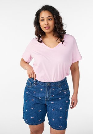 Vrouw met golvend donker haar, gekleed in een lichtroze V-hals T-shirt en hooggetailleerde denim shorts met een patroon van kleine roze strikjes.