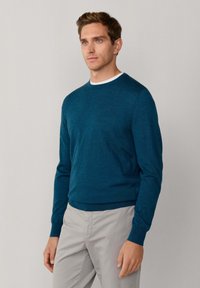 Męski sweter w kolorze ciemnozielonym, wykonany z miękkiej tkaniny. Posiada długie rękawy i subtelne logo. Połączony z jasnoszarymi spodniami.