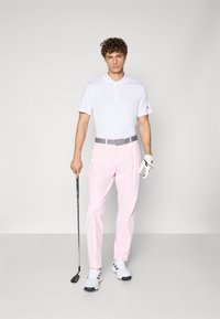 Bílá polo košile, růžové kalhoty, šedý opasek a golfová rukavice. Postoj s černým golfovým holem. Bílé sportovní boty s červenými akcenty.