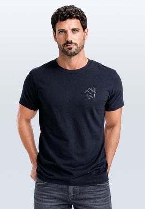 Man with short curly hair and beard wears black t-shirt with small white mountain and sun design on chest, standing against light background.