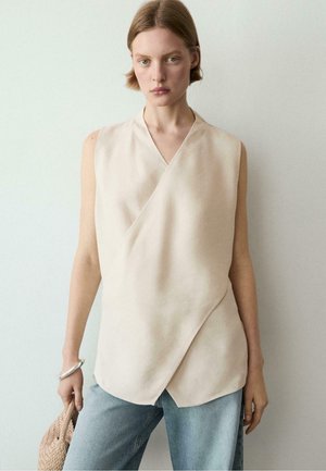 Femme aux cheveux courts portant un haut beige sans manches cache-cœur et un jean bleu, tenant un sac tressé et portant un bracelet en argent.