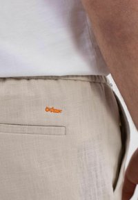 Beige Leinenhose mit elastischem Bund, versehen mit einem kleinen orangefarbenen gestickten Logo und einer Gesäßtasche. Texturiertes Material mit lockerer Passform.
