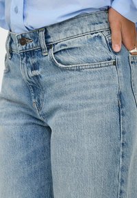 Jeans de denim azul claro con un diseño clásico de cinco bolsillos, costuras visibles y un cierre de botón en bronce. Textura ligeramente desgastada.