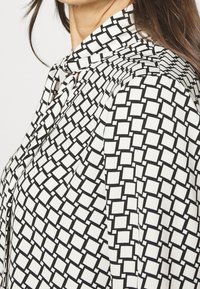 Blusa bianca e nera con un motivo geometrico a quadrati, caratterizzata da un collo a cravatta e tessuto morbido e leggero. Texture liscia e design strutturato.