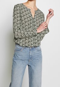Blouse à manches longues, verte avec un motif géométrique en crème et olive. Présente un col rond et un détail de boutons à l'avant. Associée à un jean bleu.