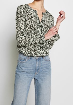 Blouse à manches longues, verte avec un motif géométrique en crème et olive. Présente un col rond et un détail de boutons à l'avant. Associée à un jean bleu.