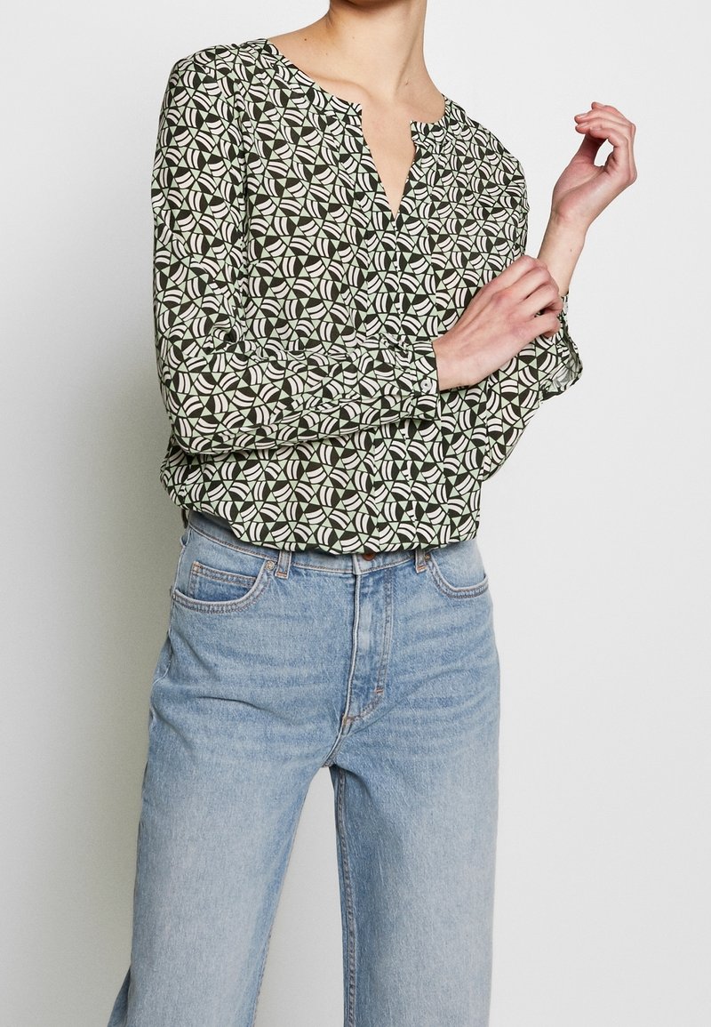 Blouse à manches longues, verte avec un motif géométrique en crème et olive. Présente un col rond et un détail de boutons à l'avant. Associée à un jean bleu.