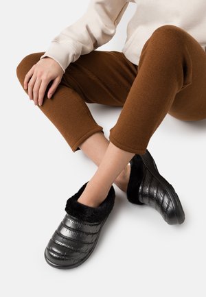 Chaussons noirs matelassés avec une doublure en fausse fourrure noire et douce, présentant un bout arrondi et une surface texturée, posés sur un pantalon marron sur fond blanc.