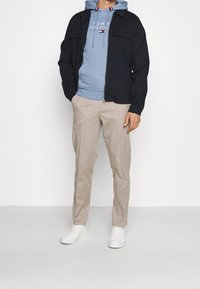 Chaqueta navy con cremallera, sudadera azul claro con logotipo, pantalones chinos beige y zapatillas blancas. La chaqueta tiene dos bolsillos frontales y un corte relajado.