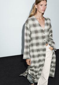 Szara i biała koszula w kratę z dekoltem w szpic, ozdobnymi guzikami i luźnymi rękawami, w połączeniu z jasnymi spodniami. Gładka tekstura materiału.
