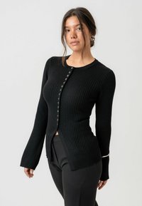 Schwarze gerippte Strickjacke mit tailliertem Schnitt, langen Ärmeln und Knopfverschluss. Hervorzuhebende Details sind ein kleiner seitlicher Schlitz.