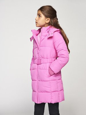 Junges Mädchen mit braunem Haar, das einen taillierten, pinken Daunenmantel trägt und mit Händen in den Taschen nach links schaut, vor einem schlichten Hintergrund.