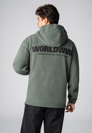 Grüner Fleece-Hoodie mit einer großen schwarzen "WORLDWIDE" Grafik auf dem Rücken. Verfügt über eine Kapuze und lange Ärmel, kombiniert mit dunklen Hosen.