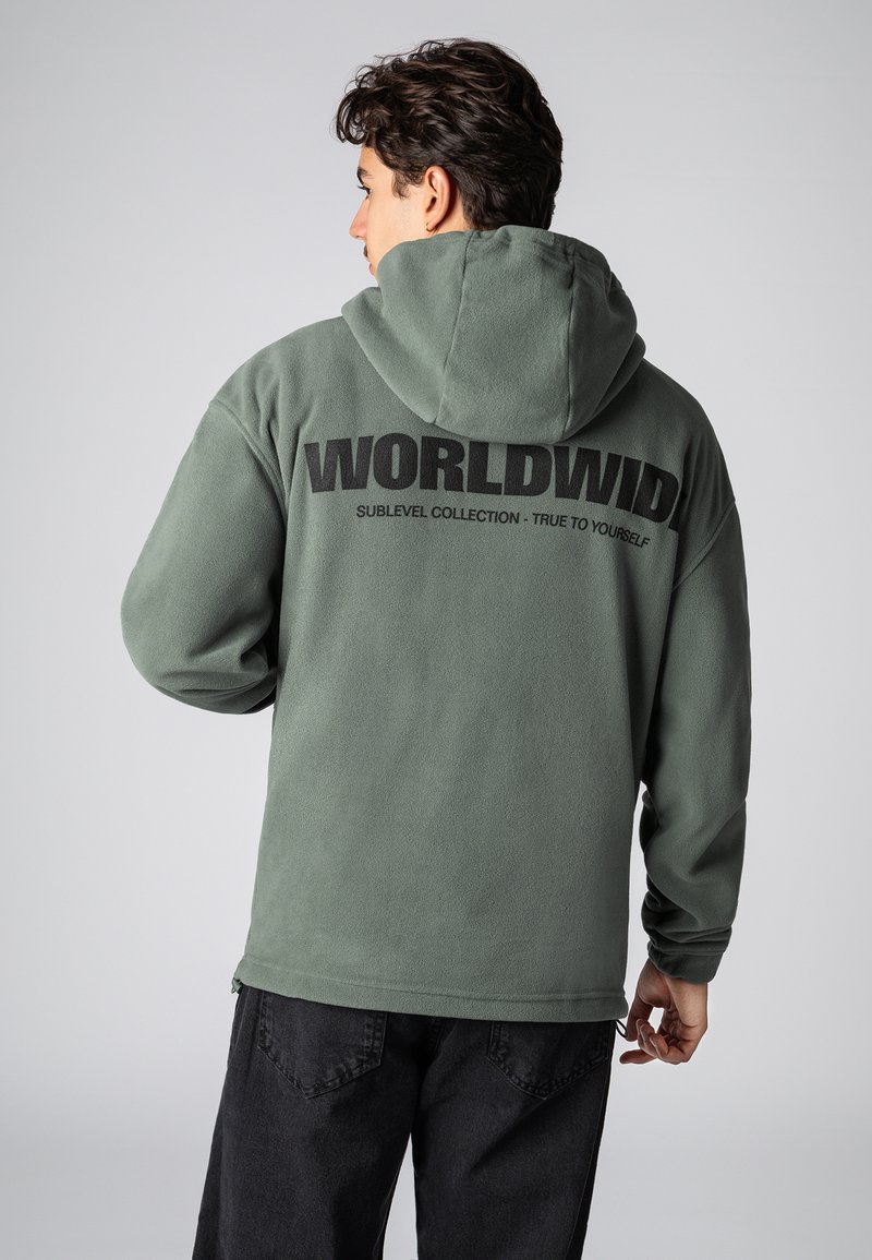 Grüner Fleece-Hoodie mit einer großen schwarzen "WORLDWIDE" Grafik auf dem Rücken. Verfügt über eine Kapuze und lange Ärmel, kombiniert mit dunklen Hosen.