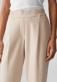 Pantaloni beige su misura con tessuto testurizzato, caratterizzati da una vita aderente e pieghe frontali, mostrati dalla vita in giù con le mani appoggiate nelle vicinanze.