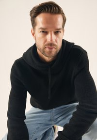 Hoodie noir en tricot avec un design bouclé, capuche attachée et cordon de serrage. Porté avec un jean en denim bleu clair. Tissu texturé visible.