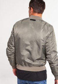 Bavlněná bunda v světle olivové barvě, vyrobená z hladkého saténového materiálu; obsahuje žebrované manžety a lem, a subtílní černou nášivku na límci.