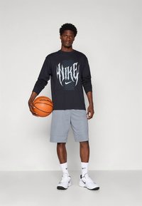 Svart langermet skjorte med "NIKE"-grafikk, grå treningsshorts, hvite joggesko, holder en oransje basketball. Ensfarget bakgrunn.