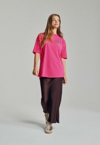 Helles pinkes Oversized-T-Shirt mit buntem Stickmuster auf der Brust, kombiniert mit einem langen braunen Satinrock und beigen sowie grauen Schnürsneakern.