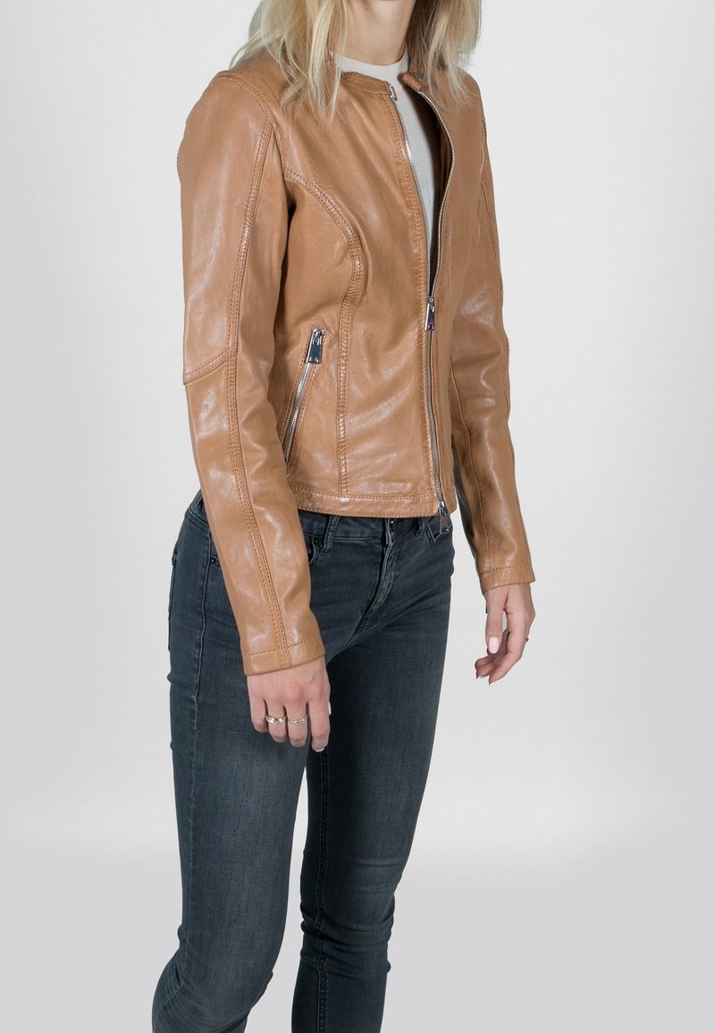 Personne portant une veste en cuir beige avec des poches zippées sur un haut blanc et un jean skinny foncé sur un fond clair uni.