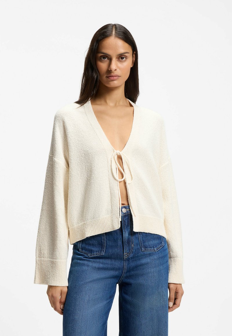 Femme aux longs cheveux foncés portant un cardigan en tricot couleur crème noué une fois au niveau de la poitrine et un jean bleu taille haute, debout devant un fond blanc.