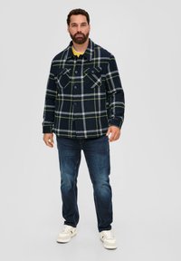 Blau kariertes Flanellhemd mit gelben Akzentlinien, ausgestattet mit zwei Brusttaschen und einer Knopfleiste, kombiniert mit dunklen Jeans und weißen Sneakers.