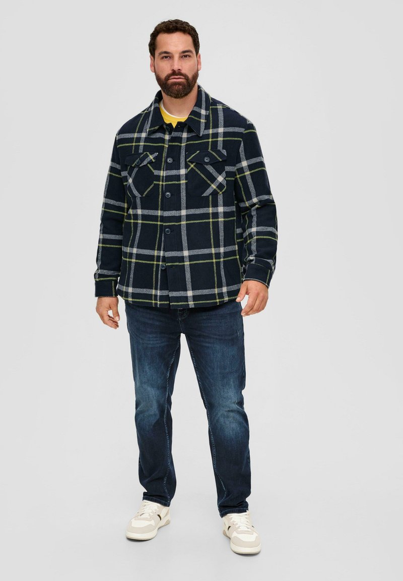 Blau kariertes Flanellhemd mit gelben Akzentlinien, ausgestattet mit zwei Brusttaschen und einer Knopfleiste, kombiniert mit dunklen Jeans und weißen Sneakers.