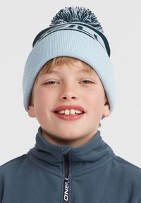 Berretto a maglia blu chiaro con una fascia scura e pon-pon sulla parte superiore. Indossato da un bambino in una giacca in pile grigia con colletto alto.