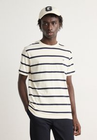 Stribet kortærmet t-shirt i creme med marineblå vandrette striber, i bomulds materiale. Båret med en beige cap, der har et logo på forsiden.