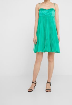 Kvinde iført en ærmeløs, plisseret smaragdgrøn kjole med tynde stropper og sorte høje hæle sandaler, stående mod en ensfarvet, lys baggrund.