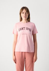 T-shirt di cotone rosa chiaro con stampa "GANT 1949 NEW HAVEN" in marrone scuro e bianco. Maniche corte, scollo rotondo, vestibilità morbida.