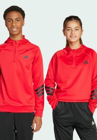 Sudaderas deportivas rojas con cremallera en el cuello, que presentan acentos a rayas negras en las mangas y el logo. Combinadas con pantalones negros. Textura de tela suave.