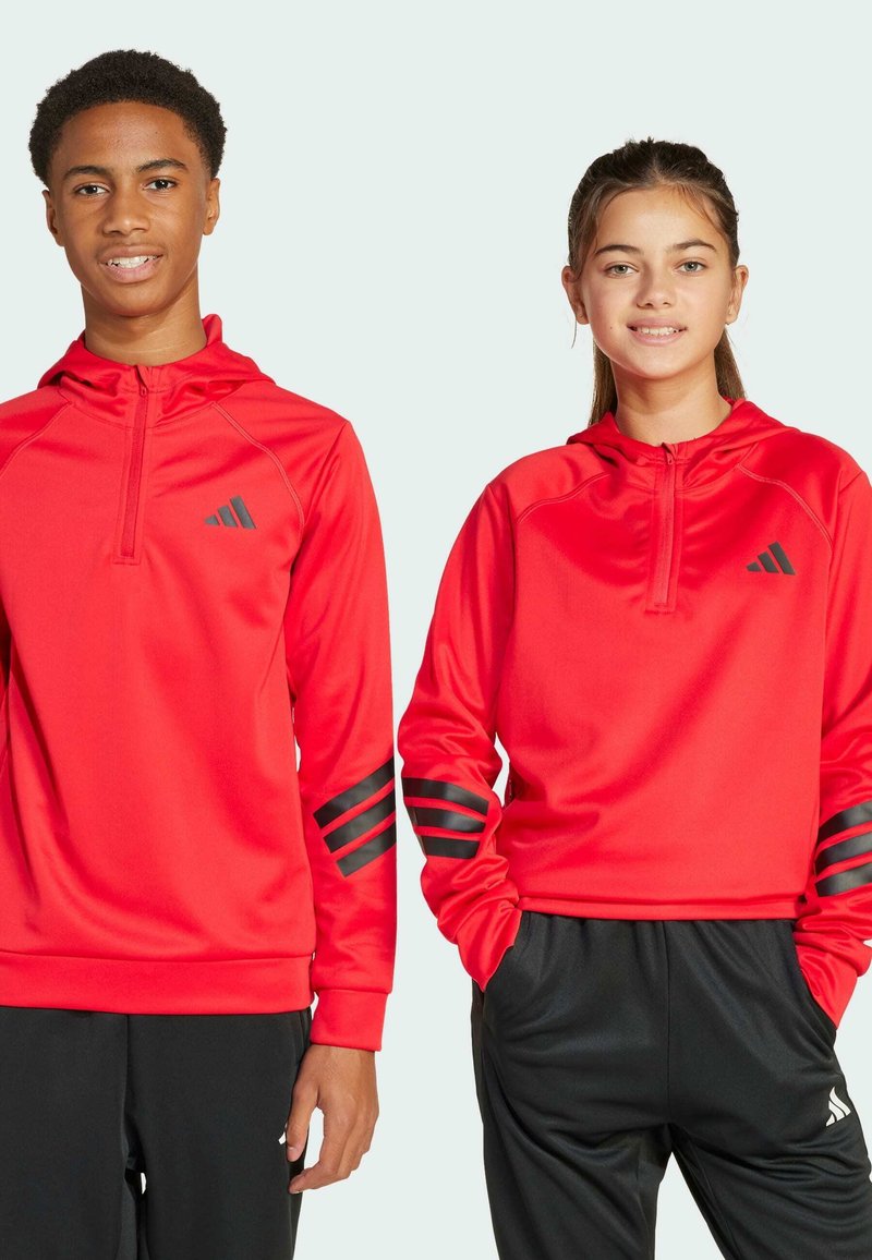 Sudaderas deportivas rojas con cremallera en el cuello, que presentan acentos a rayas negras en las mangas y el logo. Combinadas con pantalones negros. Textura de tela suave.