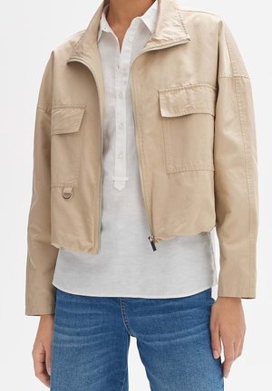 Beige cropped jakke med lynlås og klaplommer båret over en hvid skjorte med knapper og blå jeans.
