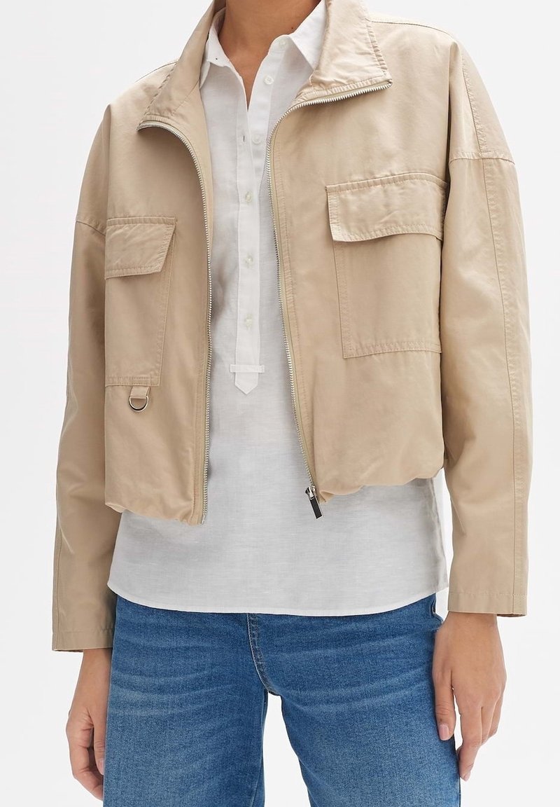 Veste beige courte avec fermeture éclair et poches à rabat portée sur une chemise blanche boutonnée et un jean bleu.