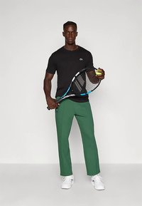 Άνδρας που στέκεται με ρακέτα τένις στο ένα χέρι και μπάλα τένις στο άλλο, φοράει μαύρο πουκάμισο, πράσινο παντελόνι και λευκά αθλητικά παπούτσια.