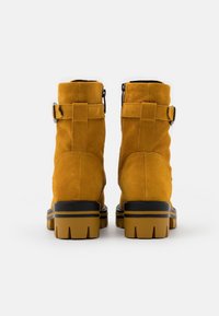 Bottines en cuir en suède jaune moutarde, avec un bout arrondi et une semelle en caoutchouc noir à rainures, maintenues par des sangles ajustables.