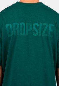 Grünes Baumwoll-T-Shirt mit einem matten, übergroßen Druck des Wortes "DROPSIZE" in fetten Buchstaben auf der Rückseite. Kurzarm und rundem Halsausschnitt.