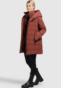Woman wearing a rust-colored quilted hooded coat, black leggings, and black ankle boots, standing against a plain light background.