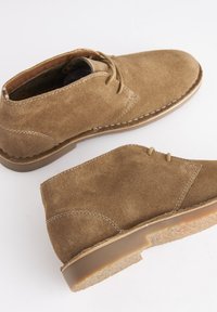 Paire de bottines desert en daim marron avec semelles en caoutchouc crêpé et fermeture à lacets, vues de côté et de dessus sur fond blanc.