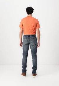 Hombre visto desde atrás con una camiseta de algodón naranja y jeans de mezclilla azules, con zapatos marrones. Los jeans presentan un diseño estándar de cinco bolsillos.