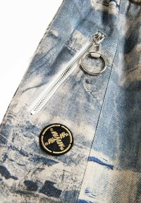 Tejido de mezclilla en azul claro con un estampado texturizado, cuenta con una cremallera plateada con tira en forma de anillo y un parche bordado negro en forma redonda.