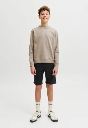 Teismeline poiss lokkis juustega, kes kannab beežikat pusa, musti lühikesi pükse, valgeid sokke ja valgeid tenniseid, seisab ühevärvilise valge tausta ees.