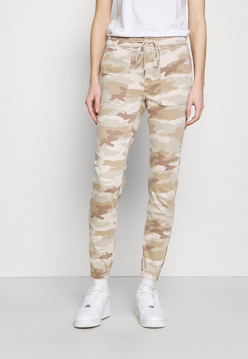 Pantalon jogger camouflage en tons beige avec taille élastique, cordon de serrage, poches latérales et chevilles resserrées. Associé à des baskets blanches.