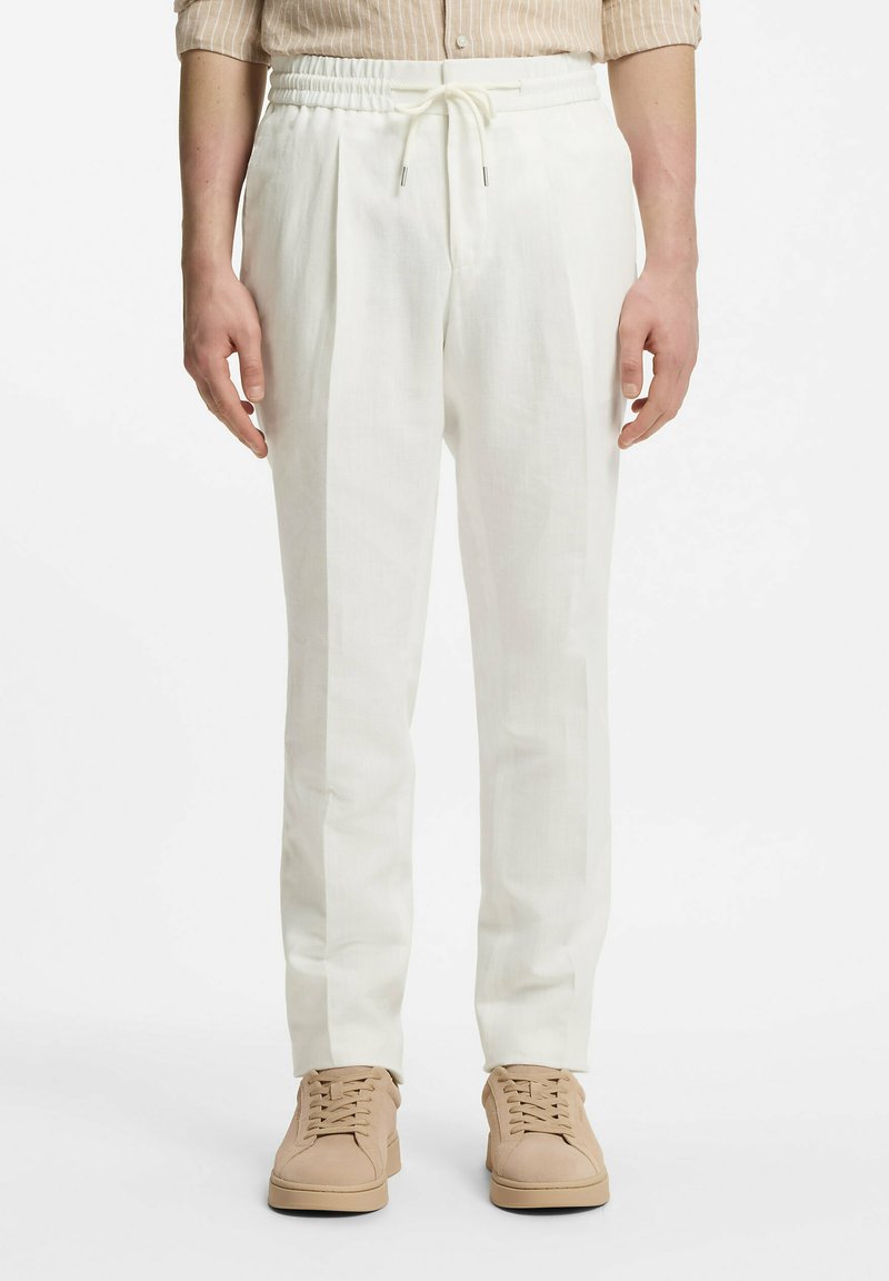 Personne portant un pantalon blanc à cordon, des baskets beiges et une chemise rayée beige, debout face à l'avant sur un fond blanc.