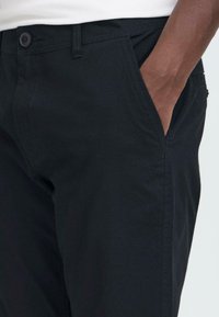 Main droite insérée dans la poche avant d'un pantalon noir porté avec une chemise blanche rentrée.