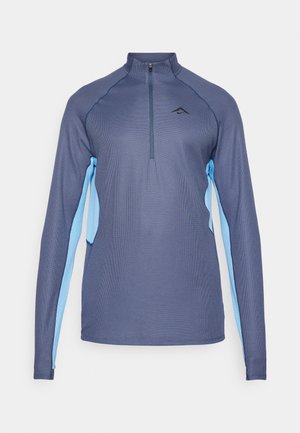 Camiseta deportiva de manga larga en azul oscuro con paneles laterales en azul claro, medio cierre de cremallera y tela texturizada. Presenta un logotipo en el pecho.